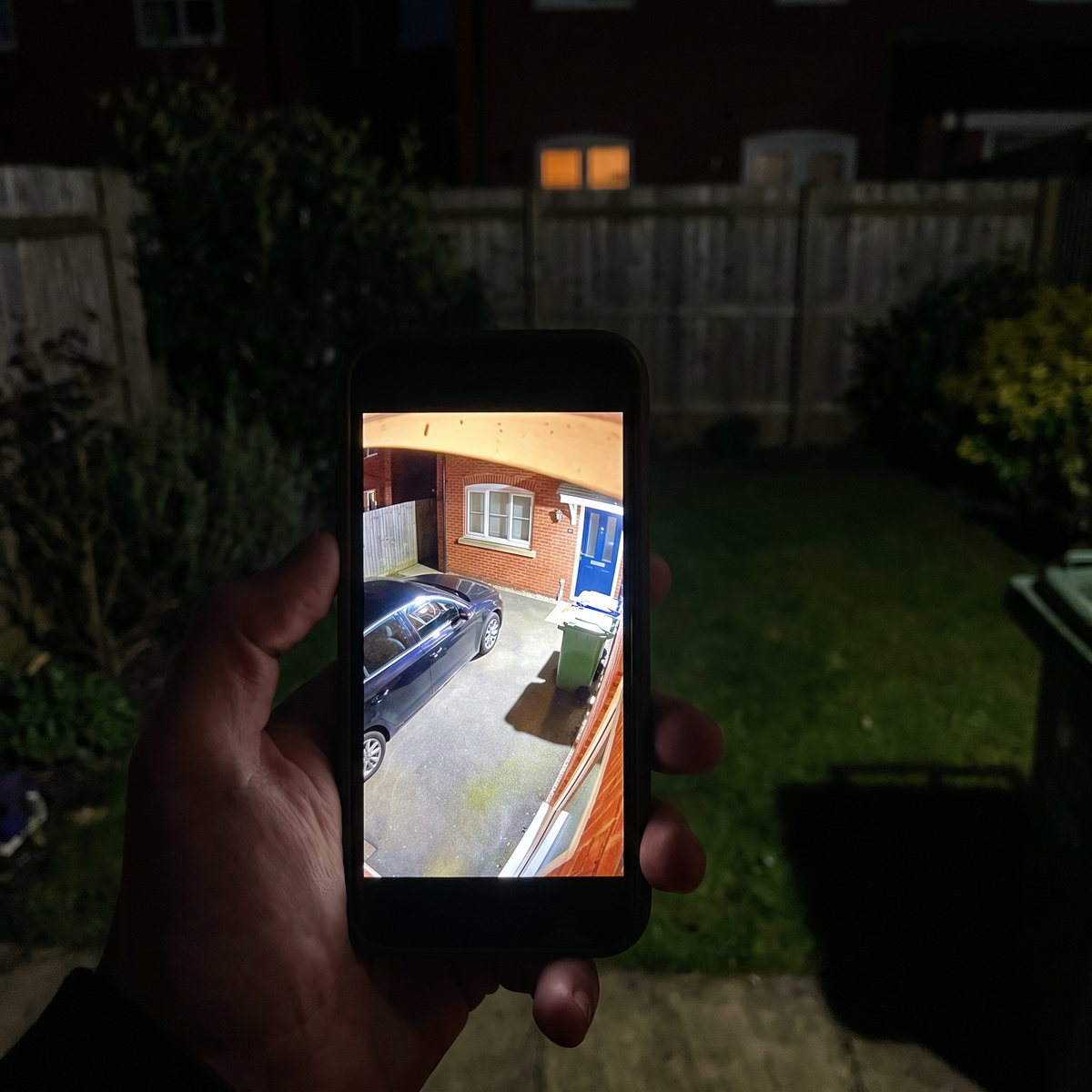 A phone held in a dark garden showing the camera's floodlit view of the driveway in full color